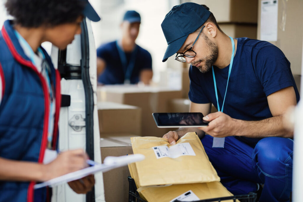 Young delivery man using digital tablet for scanning bar code on package label while preparing for shipment with his coworker.